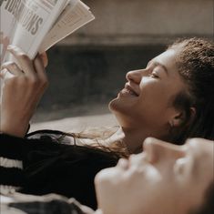 a woman laying on the ground reading a newspaper and smiling at someone else with her eyes closed