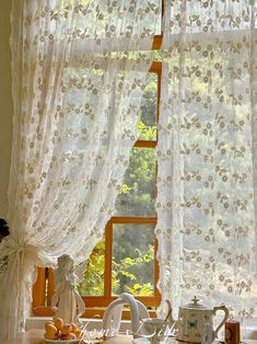 a kitchen window covered in white curtains next to a tea pot and cup on a counter