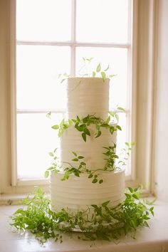 a three tiered cake with greenery on the top is displayed in front of a window