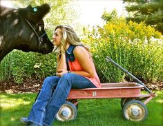 a woman sitting on top of a red wagon next to a black horse in the grass