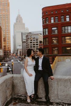 a man and woman sitting on top of a cement wall in front of tall buildings