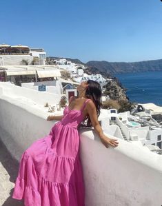 a woman in a pink dress leaning against a wall looking out at the ocean and buildings