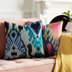 three colorful pillows on a pink couch with a plant in the corner and a potted plant next to it