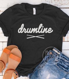 a black shirt with the word drumline on it next to denim shorts and sandals