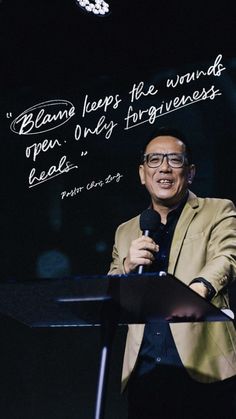 a man standing at a podium with a microphone in his hand and a quote on the wall behind him