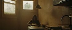 a woman sitting at a table in a kitchen writing on a piece of paper next to a window