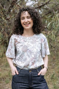 a woman with curly hair standing in front of some trees and smiling at the camera