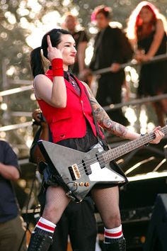 a woman in red shirt and black skirt holding a guitar on stage with other people behind her