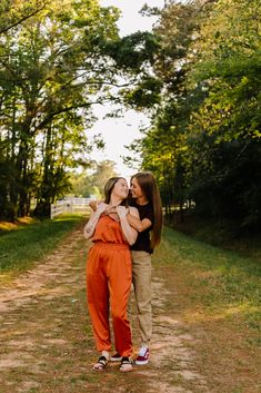 two women hugging each other in the middle of a dirt road surrounded by trees and grass