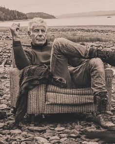 an old man sitting in a chair on some rocks by the water with his feet up