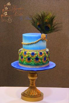 a three tiered cake with blue frosting and gold trim, decorated with peacock feathers