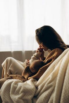 a woman sitting on a couch holding a baby and talking on a cell phone while wrapped in a blanket