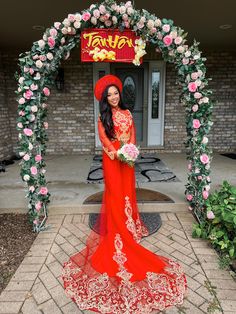 a woman in a long red dress standing under an archway with flowers on the side