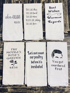 six tea towels with different sayings are on a wooden table and one is white