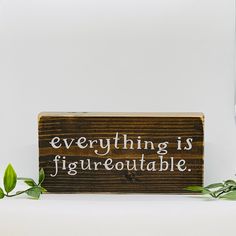 a wooden sign sitting on top of a table next to green leaves and a plant