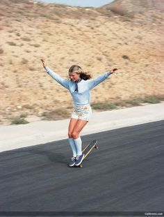 a woman riding a skateboard down a street next to a grass covered hillside and holding her arms in the air