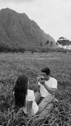 a man and woman sitting in the grass taking pictures with their cell phones while looking at each other