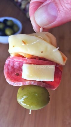 a hand holding a piece of meat and cheese on a stick with olives in the background