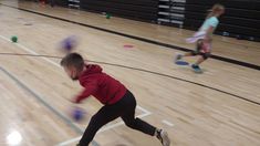 two children are playing with balls in an indoor gym flooring area while another child is running behind them