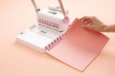 a person is using a sewing machine to sew on pink paper next to a spiral notebook