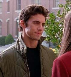 a young man standing next to a woman in front of a tree and some buildings