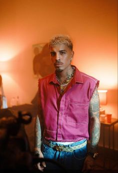 a man with tattoos wearing a pink shirt and blue jeans standing next to a lamp