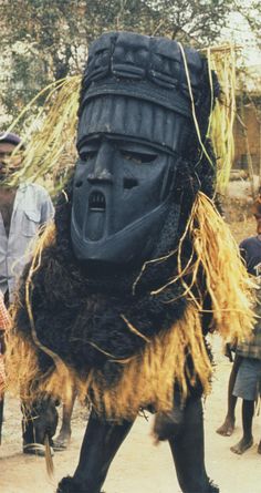 a group of people standing around a large black mask on top of a dirt ground