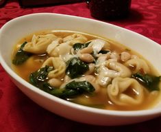 a white bowl filled with pasta and spinach in broth, on top of a red table cloth