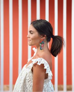 a woman in white dress standing next to a red wall and wearing large colorful earrings