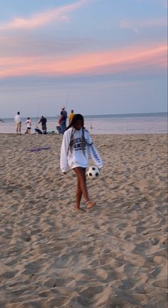 a woman walking across a sandy beach holding a soccer ball