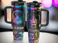 two travel mugs with designs on them sitting on a table in front of a blurry background