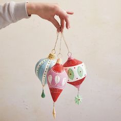 a hand holding three ornaments in different colors and shapes, hanging from strings on a wall