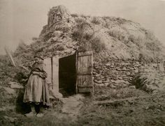 an old black and white photo of a woman standing in front of a small hut