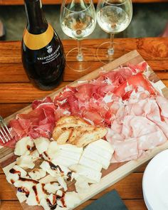 a wooden cutting board topped with meat and cheese next to two wine glasses on top of a table