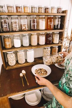 a woman standing in front of a wooden shelf filled with jars and bowls full of food