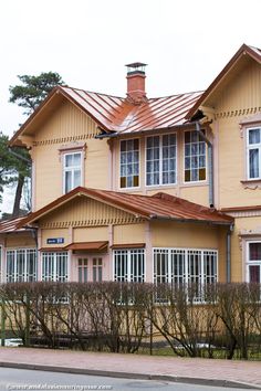 a large yellow house with white windows and a red roof on a street in front of some bushes