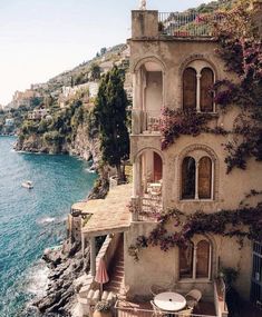 an old house on the edge of a cliff by the ocean with flowers growing all over it