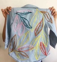 a person holding up a jean jacket with embroidered leaves on it