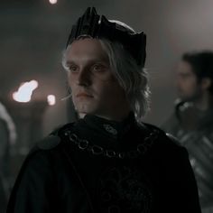 a man with white hair wearing a crown in a dark room next to other men