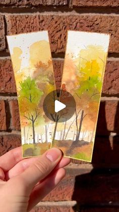 someone holding up two small cards with trees on them in front of a brick wall