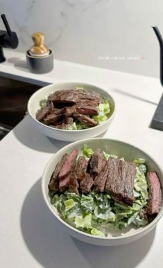 two white bowls filled with steak and lettuce on top of a countertop