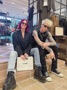 two people sitting on the steps in front of clothing racks and bags, one with tattoos