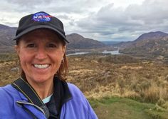 a woman wearing a blue jacket and hat standing in front of mountains with a lake behind her