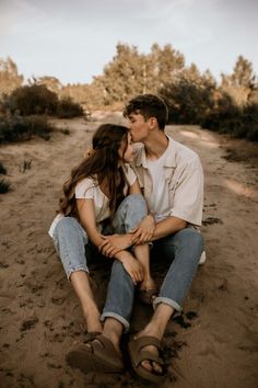 a man and woman sitting in the sand kissing each other with trees in the background