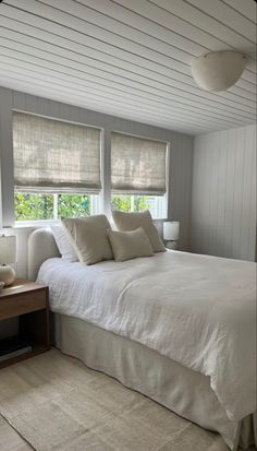 a white bed sitting in a bedroom next to two windows with roman shades on them