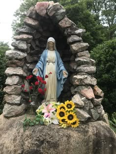 a statue of the virgin mary surrounded by sunflowers and other flowers on a rock