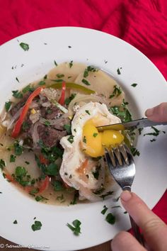 a person is holding a fork and spoon over a bowl of food on a white plate