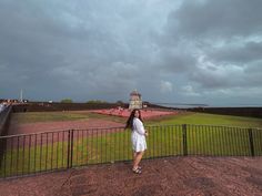 a woman standing on top of a lush green field next to a metal fence with a clock tower in the background