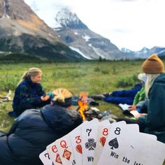 three people sitting on the ground with playing cards in front of them and mountains behind them