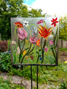 a sign with flowers painted on it sitting in the middle of a garden next to trees